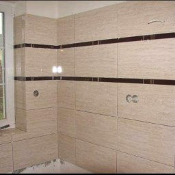 Dar-Bud - Wykończona łazienka z beżowymi płytkami imitującymi trawertyn, z ciemnobrązowymi pasami dekoracyjnymi, widoczny otwór na baterię, na parapecie leży pędzel malarski, za oknem widok na dom.