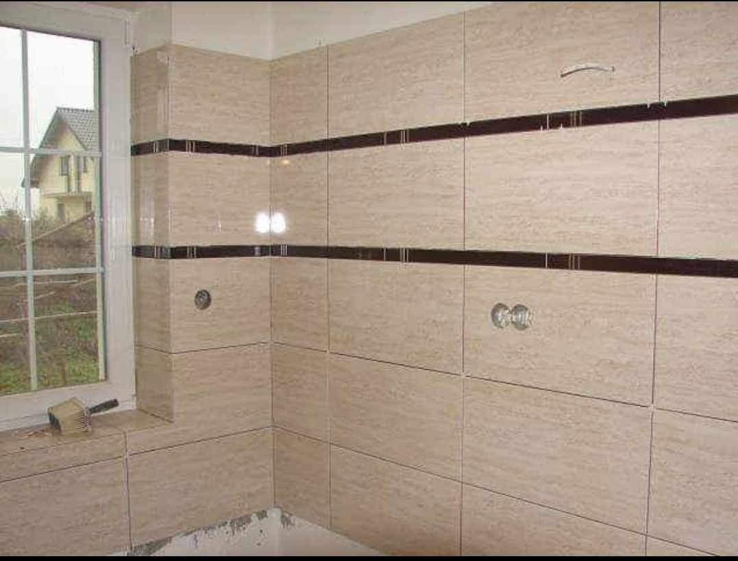 Wykończona łazienka z beżowymi płytkami imitującymi trawertyn, z ciemnobrązowymi pasami dekoracyjnymi, widoczny otwór na baterię, na parapecie leży pędzel malarski, za oknem widok na dom.