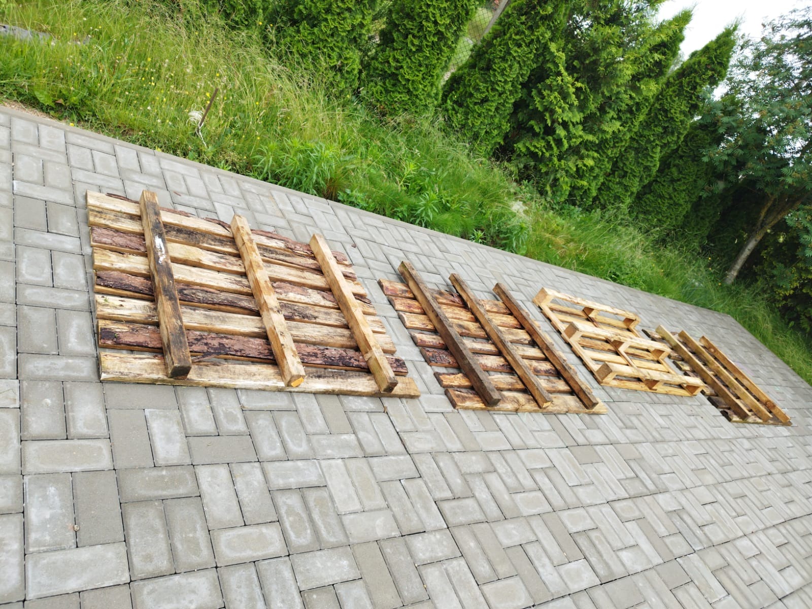 Drewniane palety ułożone na szarej kostce brukowej na zewnątrz, w tle zielone tuje i trawa.