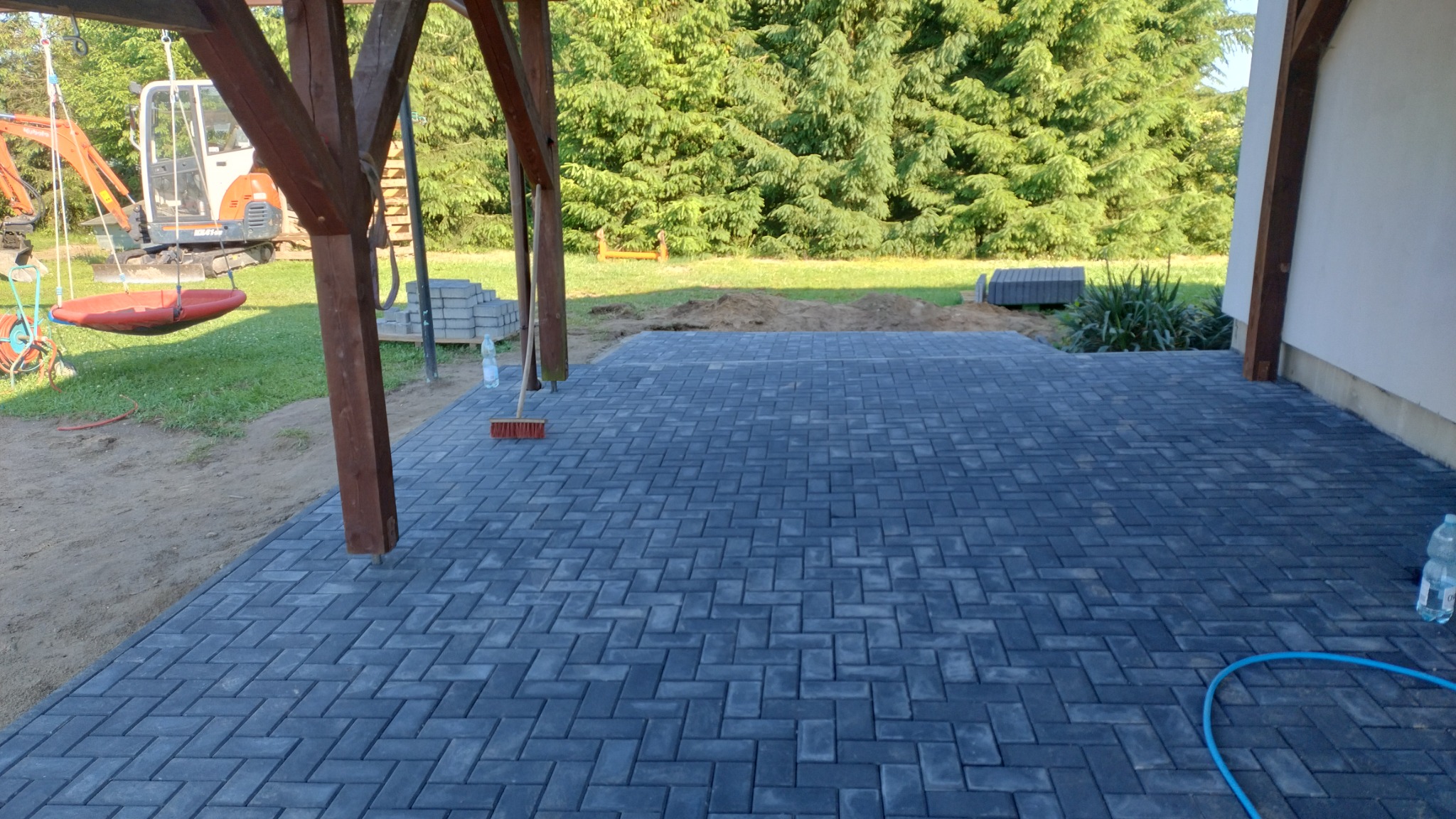 Ułożona kostka brukowa w jodełkę na tarasie z drewnianą konstrukcją wspornikową, widoczny fragment trawnika, maszyna budowlana i huśtawka w tle.