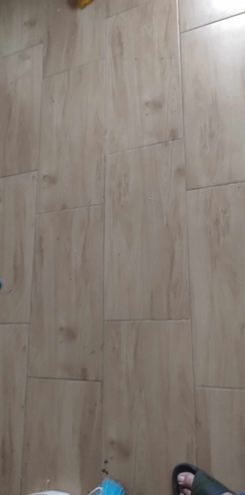 Ułożone panele podłogowe o wzorze drewna, widoczne fragment stopy w klapku i narzędzia do sprzątania w rogu.