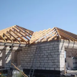 Budowa domu jednorodzinnego: ściany z białego bloczka, drewniana konstrukcja dachu w trakcie montażu, drabina oparta o ścianę, pracownik w tle.