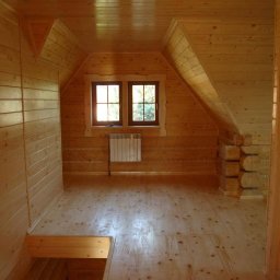 Robert Oporski Usługi Stolarsko-Budowlane - Drewniane wnętrze poddasza z oknami, wykończone boazerią, z widocznym grzejnikiem i elementami konstrukcyjnymi z bali.