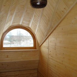 Robert Oporski Usługi Stolarsko-Budowlane - Poddasze wykończone jasnym drewnem, z oknem w kształcie łuku, przez które widać zimowy krajobraz. Na suficie widoczna dekoracyjna lampa.