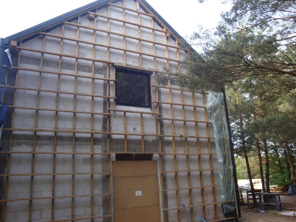 Ściana budynku w trakcie prac przygotowawczych do ocieplenia, widoczna konstrukcja drewniana przytwierdzona do muru z betonu komórkowego, okno i drzwi zabezpieczone, częściowo widoczna folia ochronna.