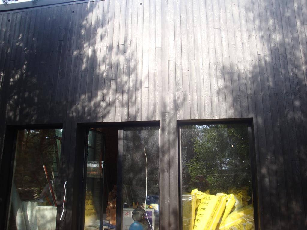 Ciemna drewniana elewacja budynku z trzema oknami, na której widoczne są cienie drzew; jedno okno jest otwarte, a wewnątrz widać materiały budowlane.