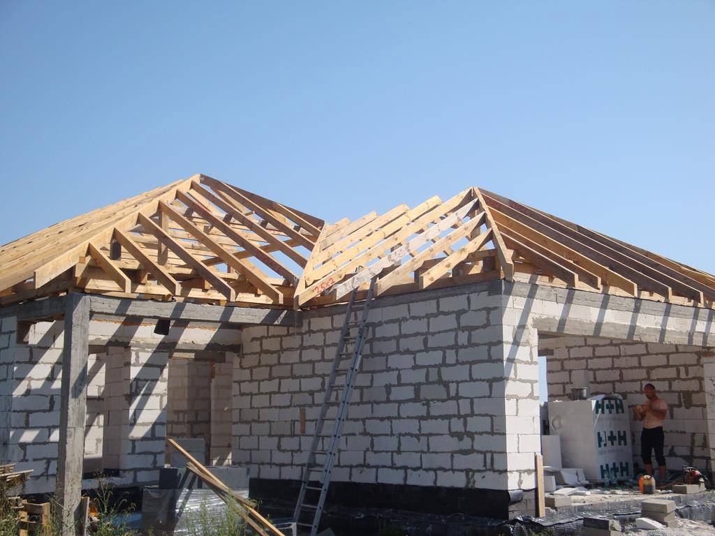 Budowa domu jednorodzinnego: ściany z białego bloczka, drewniana konstrukcja dachu w trakcie montażu, drabina oparta o ścianę, pracownik w tle.