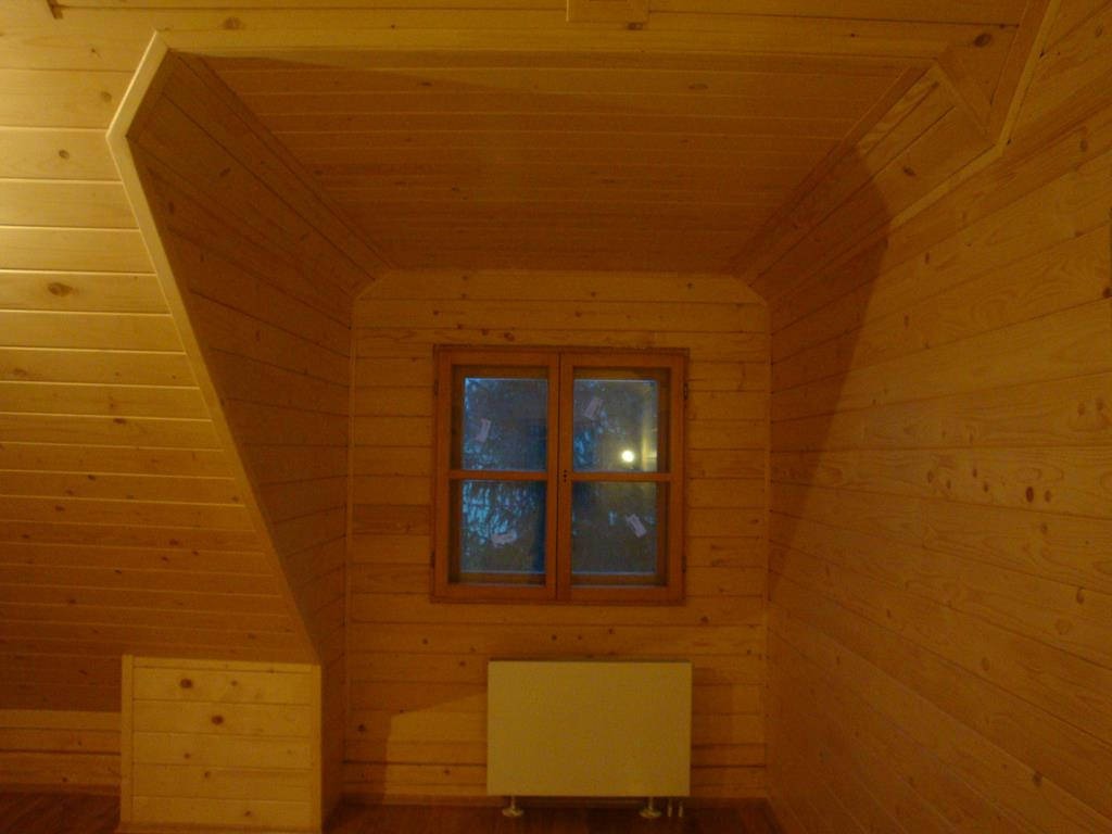 Poddasze wykończone boazerią z jasnego drewna, z oknem w drewnianej ramie i grzejnikiem pod nim.