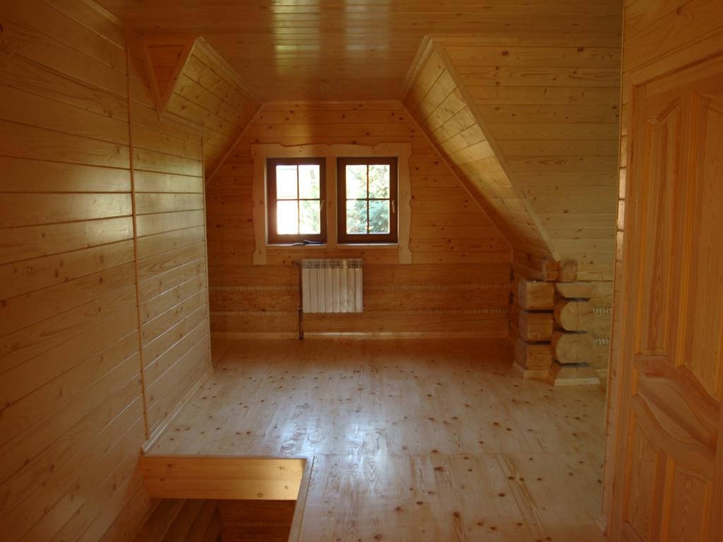 Drewniane wnętrze poddasza z oknami, wykończone boazerią, z widocznym grzejnikiem i elementami konstrukcyjnymi z bali.