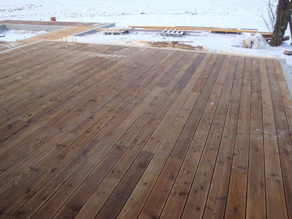 Drewniany taras z desek ryflowanych, częściowo pokryty śniegiem, z widocznymi elementami fundamentów i przygotowanymi materiałami budowlanymi w tle.