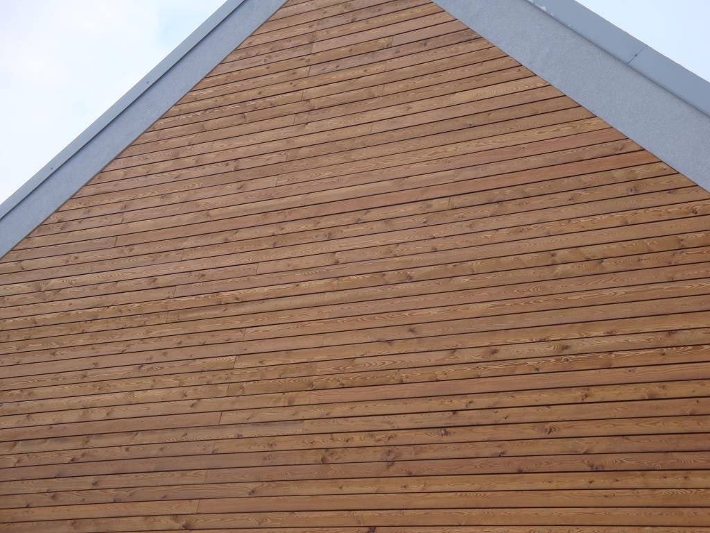 Fragment elewacji budynku z drewnianą okładziną ułożoną poziomo, widoczne szare obramowanie szczytu dachu.