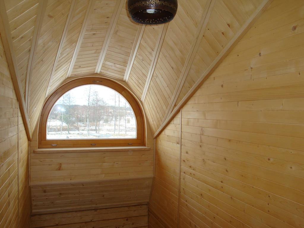 Poddasze wykończone jasnym drewnem, z oknem w kształcie łuku, przez które widać zimowy krajobraz. Na suficie widoczna dekoracyjna lampa.