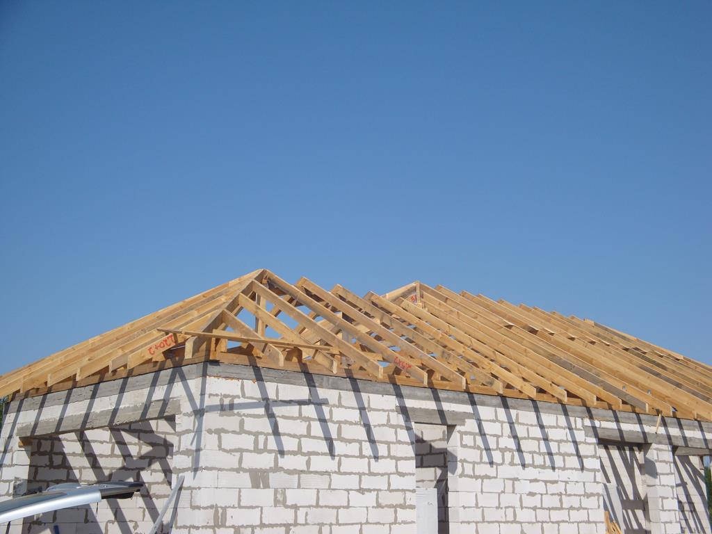 Konstrukcja drewniana dachu w trakcie budowy, widok na szkielet z belek na tle błękitnego nieba, ściany budynku z białego betonu komórkowego.