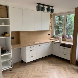 O.R. Elektro - Częściowo zmontowana kuchnia z białymi szafkami i drewnianym blatem, widoczne narzędzia i materiały wykończeniowe na blacie przy oknie, lampy sufitowe w folii ochronnej.