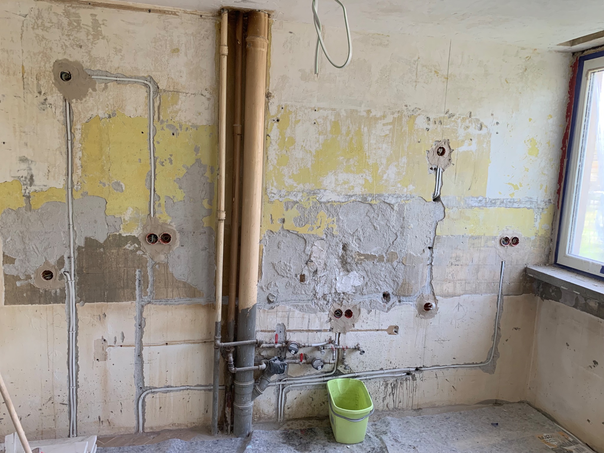 Wnętrze w trakcie remontu we Wrocławiu, widoczne poprowadzone przewody elektryczne w ścianach z przygotowanymi otworami pod gniazdka, stare rury i zielone wiadro.