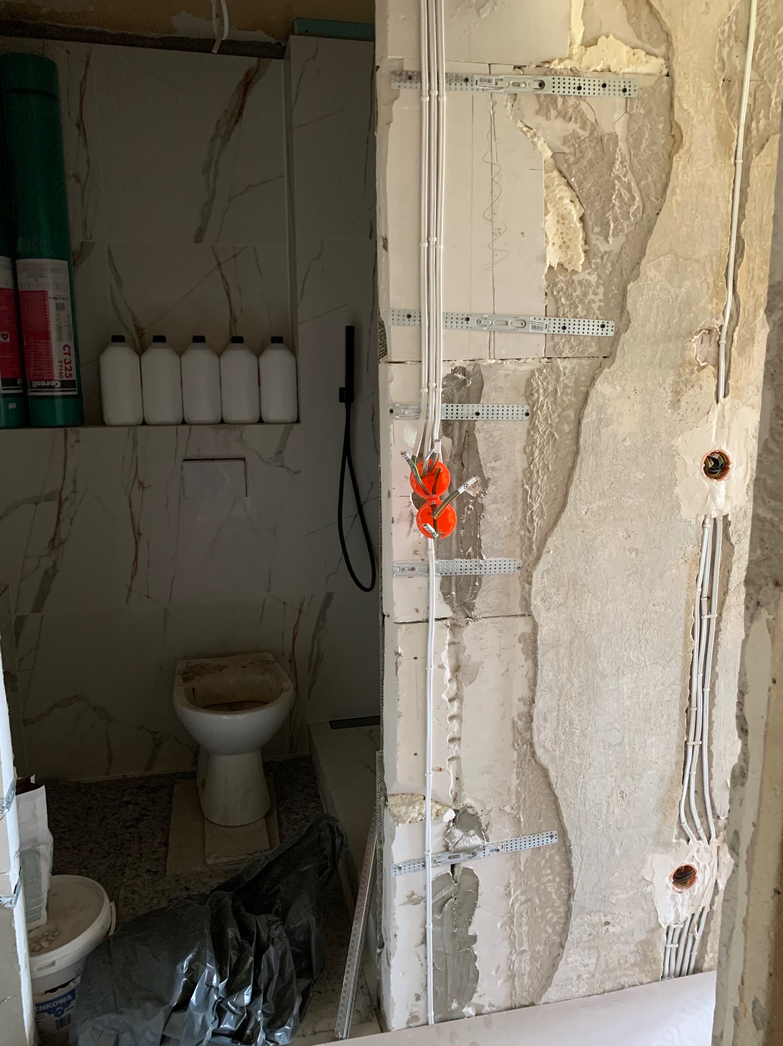 Wnętrze łazienki w trakcie remontu z widocznymi przewodami elektrycznymi w ścianie, zamontowanymi na metalowych uchwytach, oraz puszkami instalacyjnymi i częściowo wykończoną toaletą w tle.