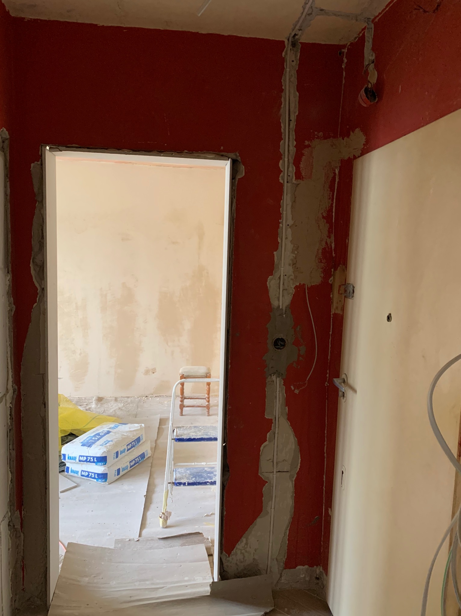 Czerwona ściana z wyżłobieniami na przewody elektryczne, widoczne kable i puszka, w tle białe worki z gipsem i drabina.