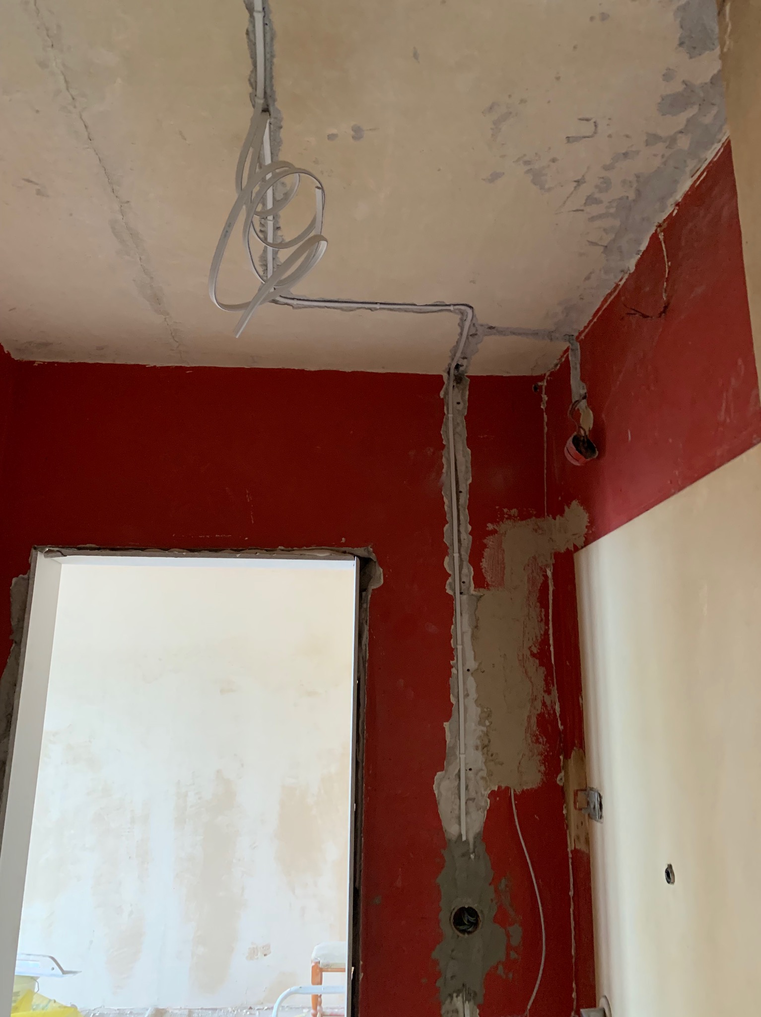 Wnętrze w trakcie remontu z widocznymi przewodami elektrycznymi poprowadzonymi po ścianie i suficie, zabezpieczonymi listwami, ściany pomalowane na czerwono i kremowo, surowy betonowy sufit.