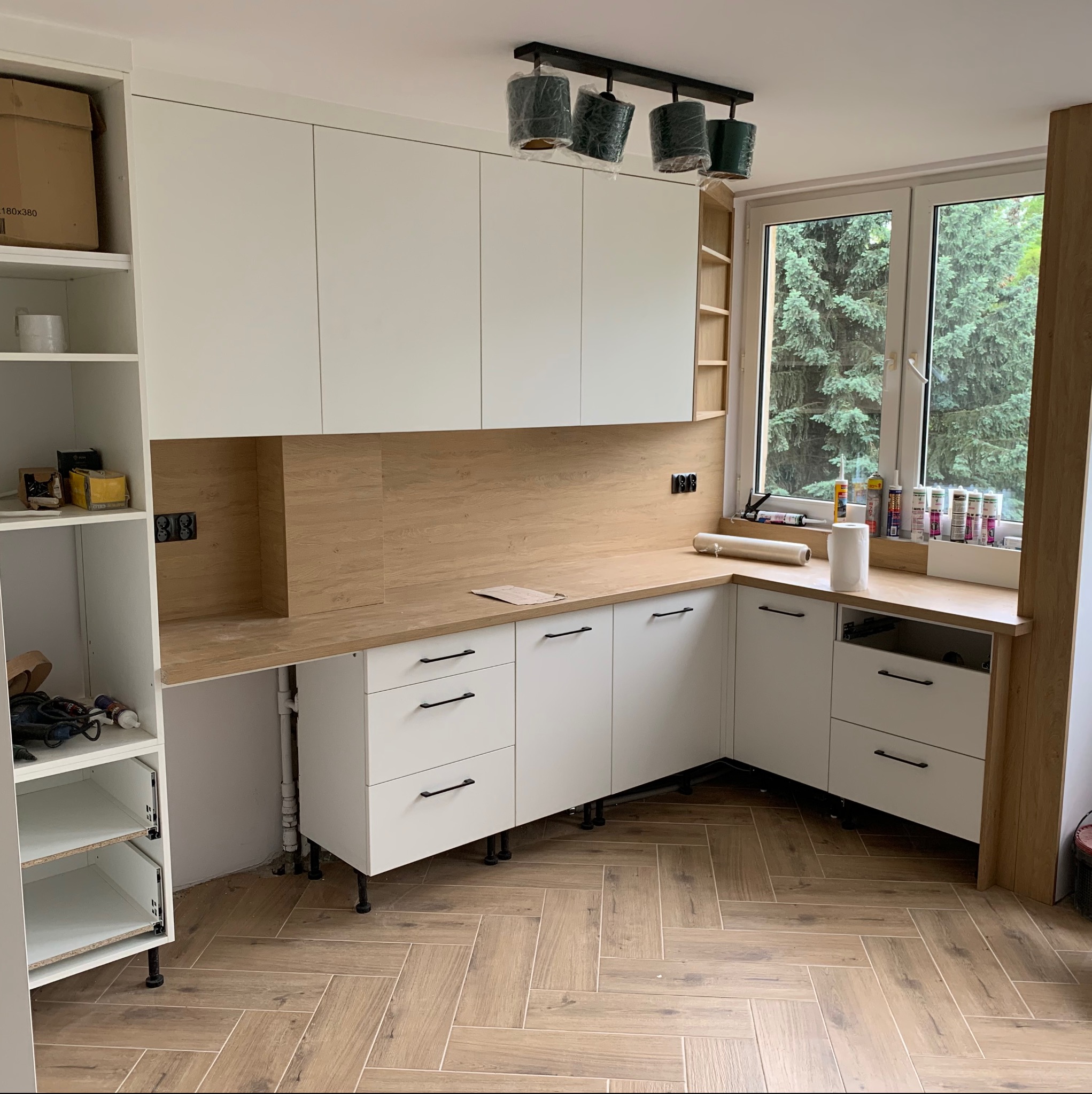 Częściowo zmontowana kuchnia z białymi szafkami i drewnianym blatem, widoczne narzędzia i materiały wykończeniowe na blacie przy oknie, lampy sufitowe w folii ochronnej.