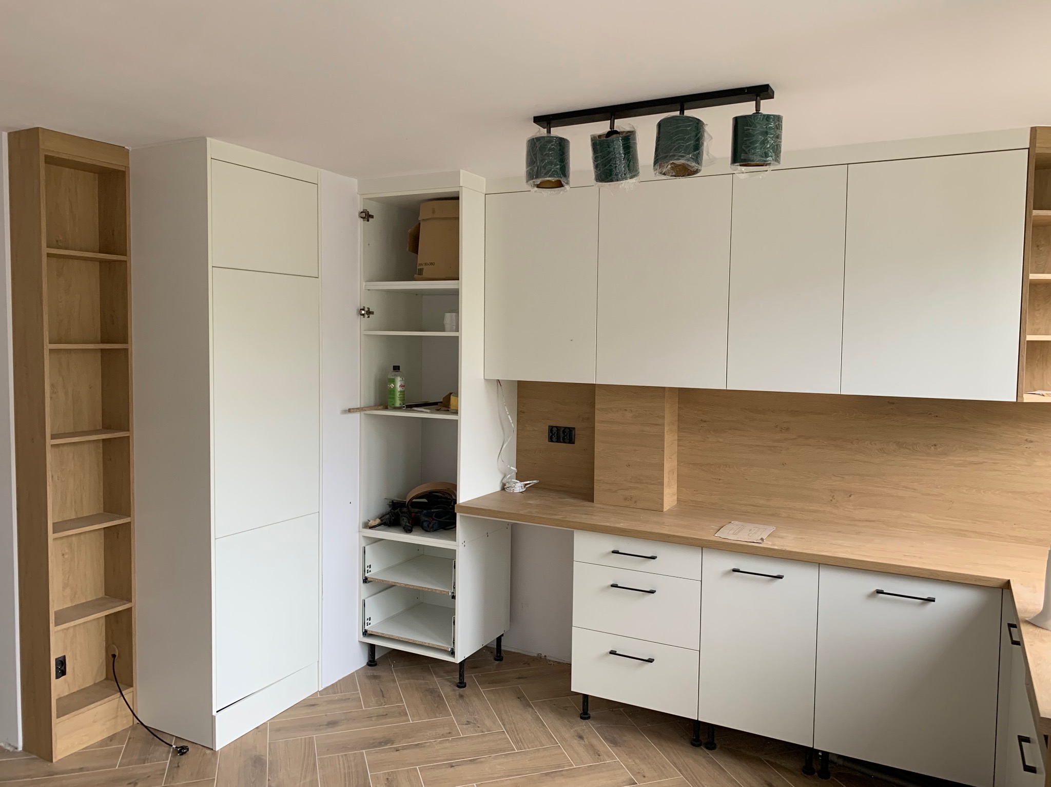 Nowoczesna, jasna kuchnia w trakcie montażu: białe szafki z drewnianym blatem i czarnymi uchwytami, otwarty słupek z półkami, lampa wisząca z trzema kloszami w folii ochronnej.