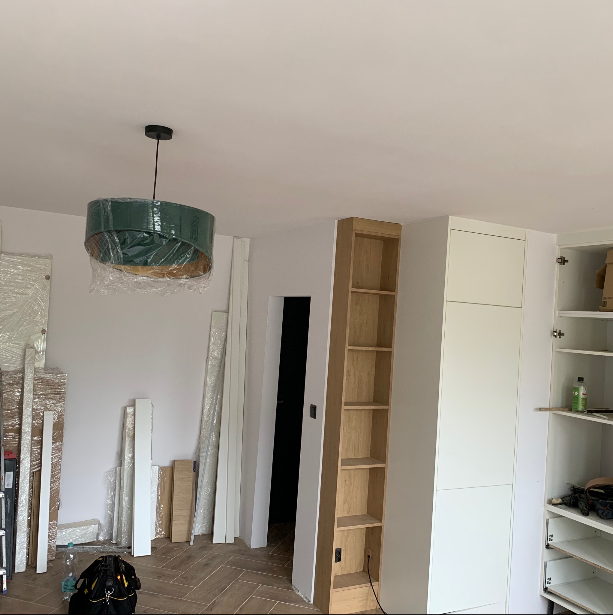 Wnętrze w trakcie wykańczania: zawieszona lampa z zielonym abażurem w folii ochronnej, drewniany regał z ukrytymi gniazdkami, biała szafa z otwartymi drzwiami ukazującymi narzędzia i środki...