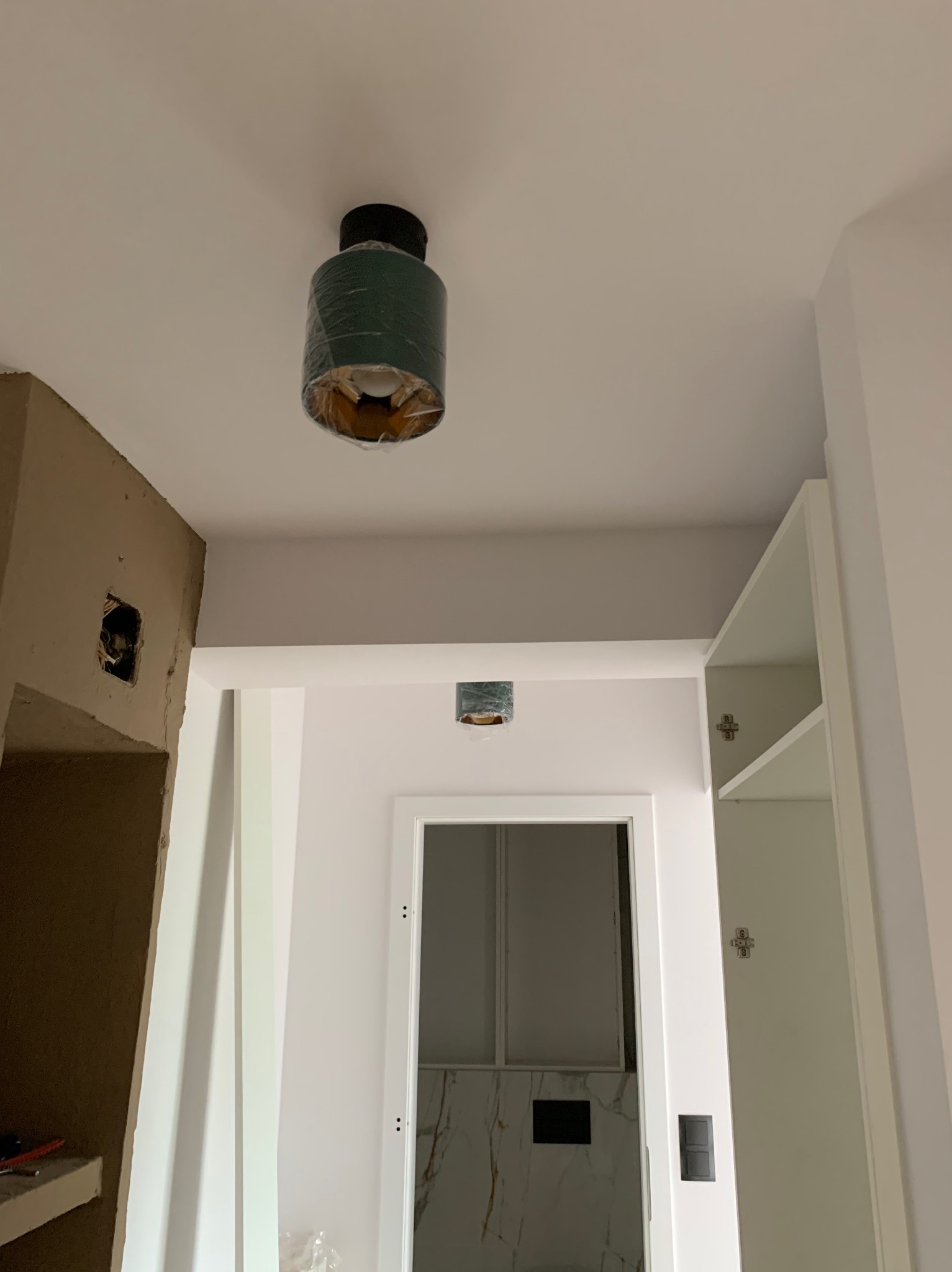 Nowa, zielona lampa sufitowa w folii ochronnej, zawieszona w pomieszczeniu w trakcie remontu, widoczna instalacja elektryczna w ścianie i otwarta szafka w tle.