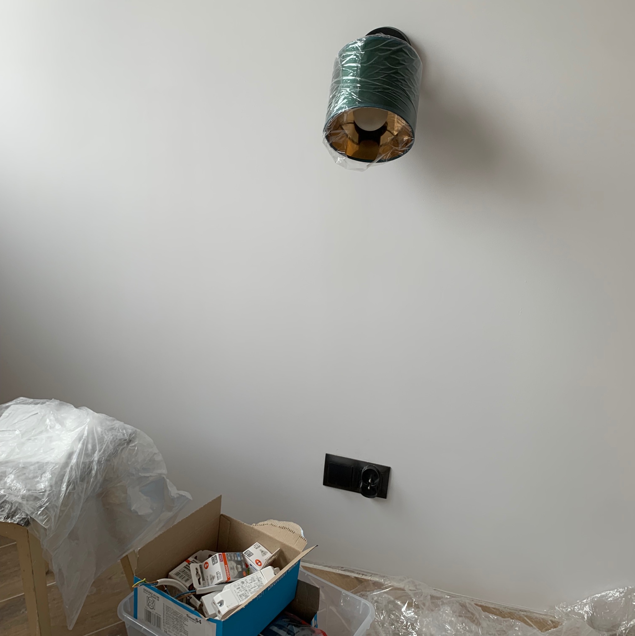 Nowoczesna, zielona lampa sufitowa z elementami w kolorze złota, jeszcze w folii ochronnej, zamontowana na białym suficie. W tle biała ściana z czarnym gniazdkiem elektrycznym i karton z elementami...