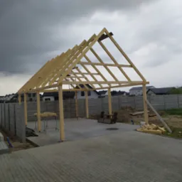 Konstrukcja drewniana z belek, prawdopodobnie zadaszenie lub wiata, w trakcie budowy, na betonowej wylewce, z widocznymi narzędziami i materiałami budowlanymi, w tle domy jednorodzinne i pochmurne...