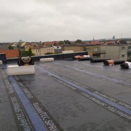Dachfix kompleksowe usługi dekarskie - Płaski dach w trakcie hydroizolacji z widocznymi rolkami papy BauderTEC Sprint DUO, bloczkami styropianowymi i radiem budowlanym DeWalt na tle zabudowy miejskiej w pochmurny dzień.