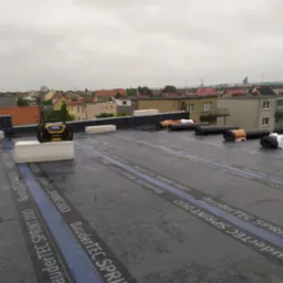 Płaski dach w trakcie hydroizolacji z widocznymi rolkami papy BauderTEC Sprint DUO, bloczkami styropianowymi i radiem budowlanym DeWalt na tle zabudowy miejskiej w pochmurny dzień.