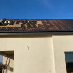 Montaż dachówki ceramicznej na nowym budynku w trakcie budowy, widoczna drabina oparta o ścianę, część dachu pokryta, a część przygotowana do położenia dachówki, błękitne niebo w tle.