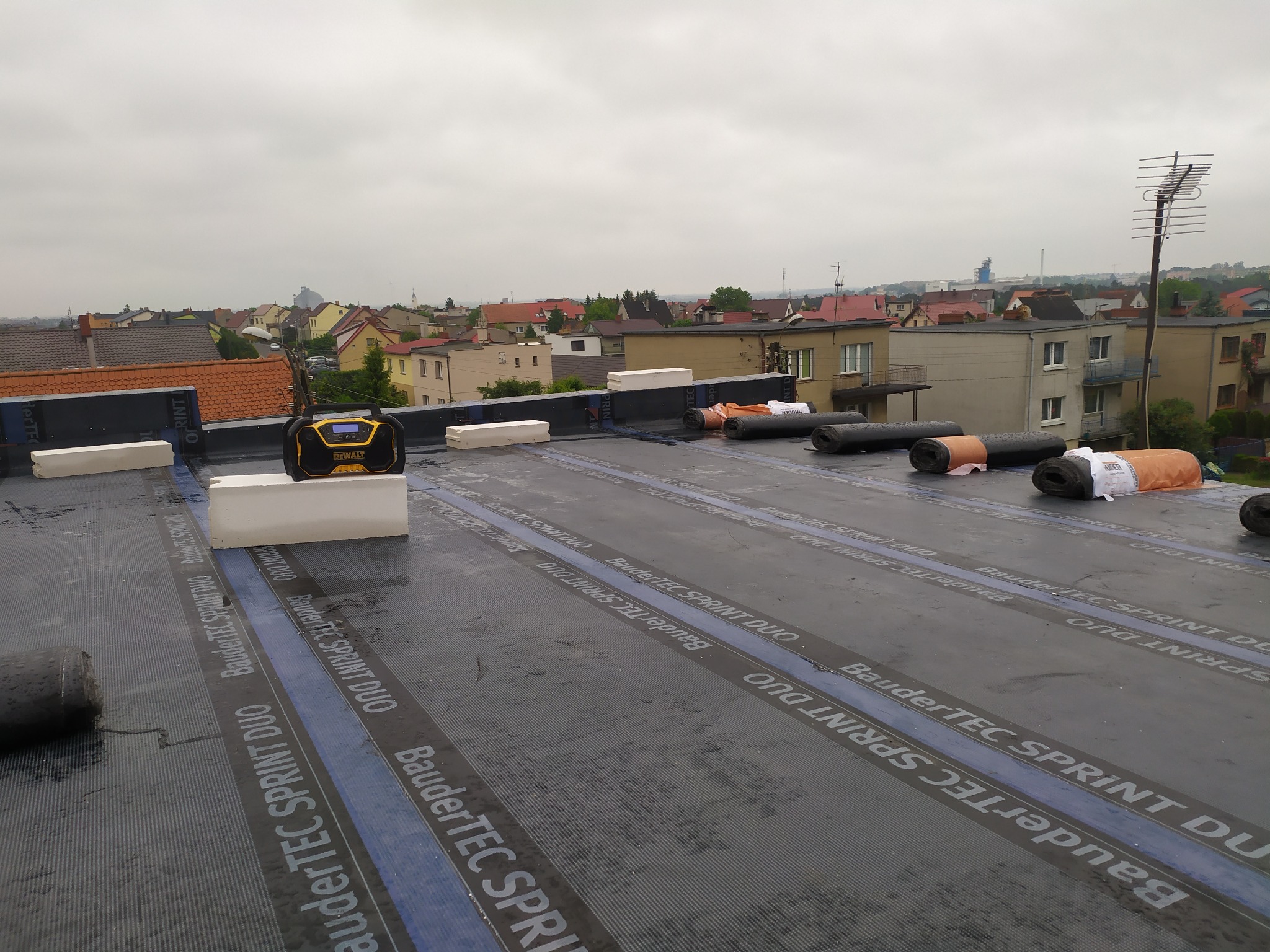 Płaski dach w trakcie hydroizolacji z widocznymi rolkami papy BauderTEC Sprint DUO, bloczkami styropianowymi i radiem budowlanym DeWalt na tle zabudowy miejskiej w pochmurny dzień.