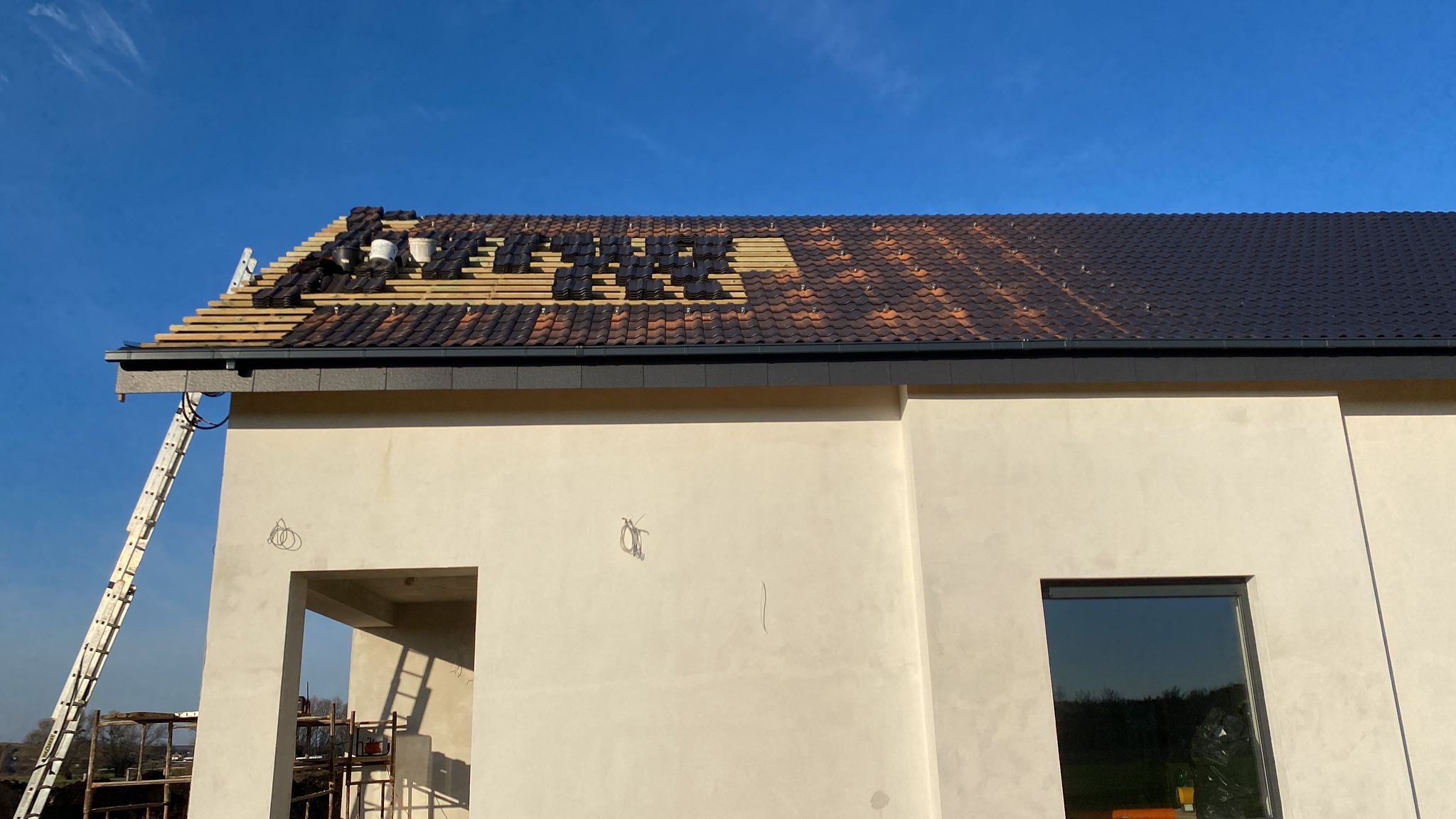 Montaż dachówki ceramicznej na nowym budynku w trakcie budowy, widoczna drabina oparta o ścianę, część dachu pokryta, a część przygotowana do położenia dachówki, błękitne niebo w tle.