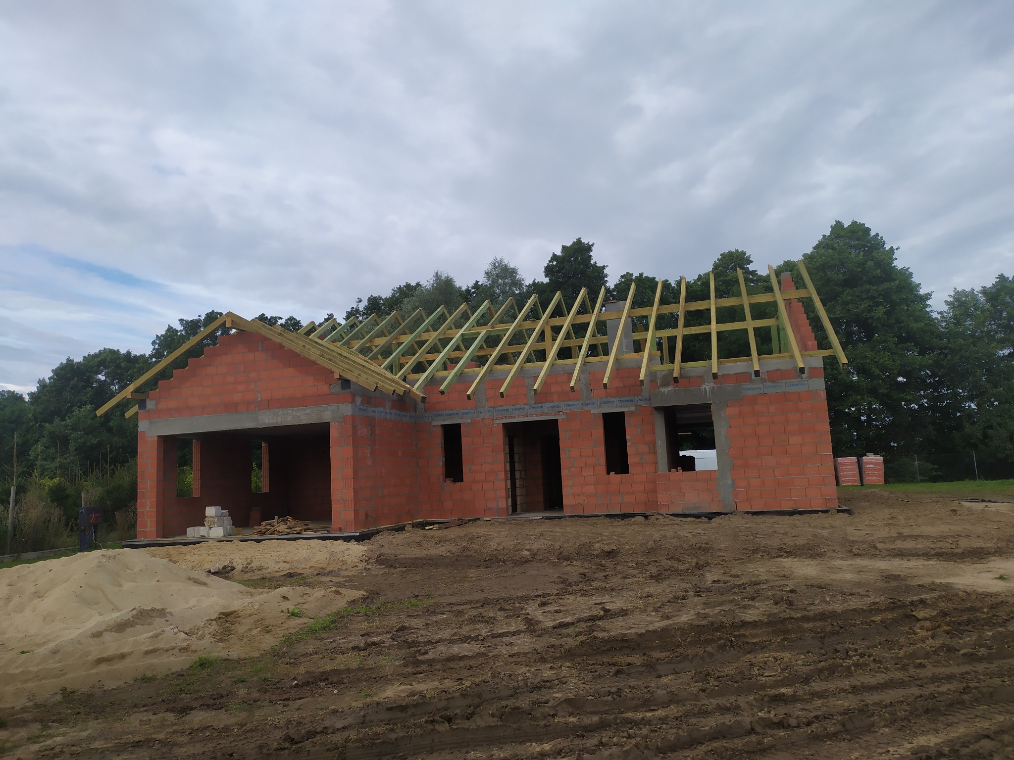 Surowy budynek jednorodzinny z czerwonej cegły z widoczną konstrukcją drewnianą dachu, otoczony ziemią i drzewami, pod pochmurnym niebem.