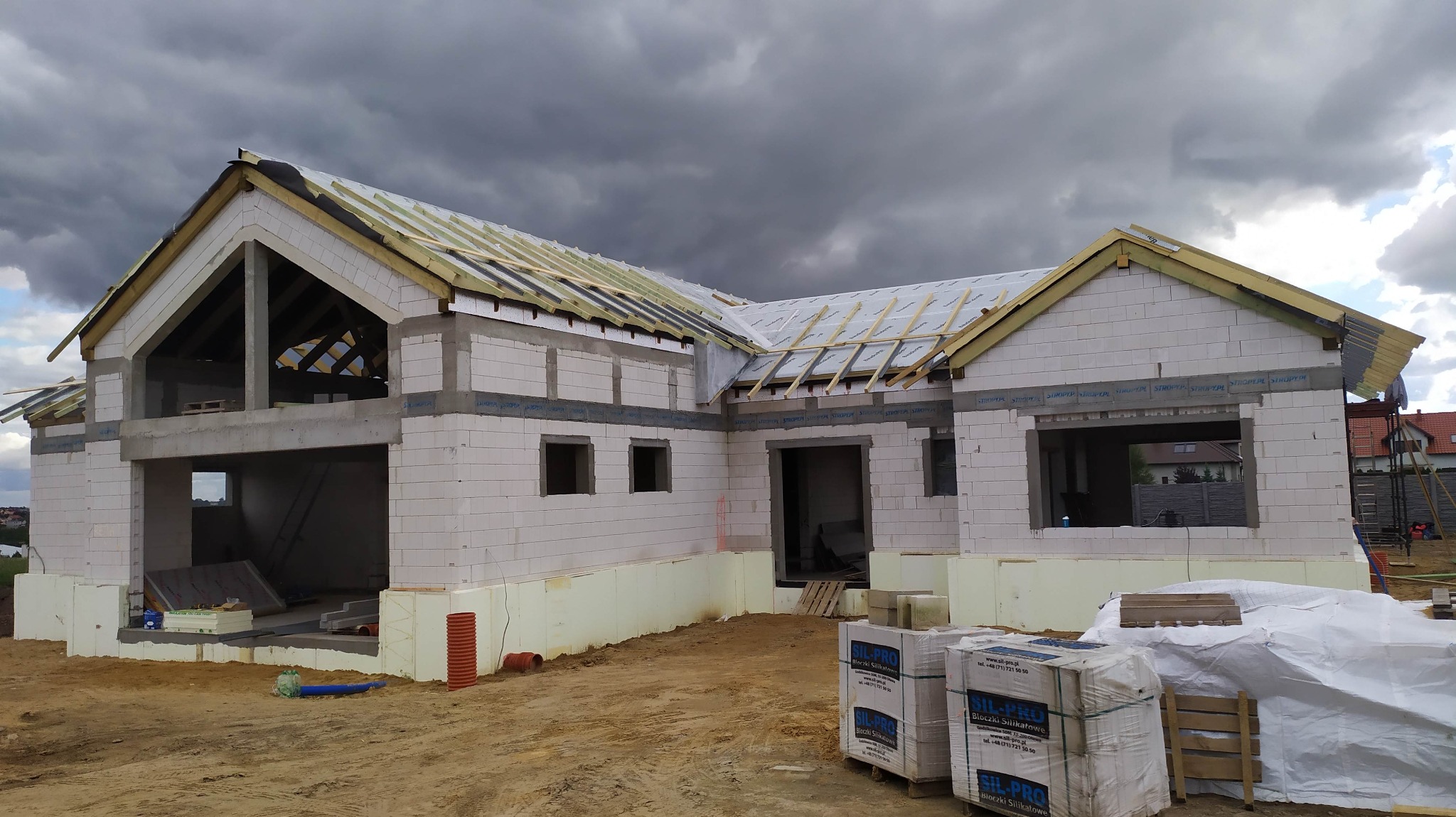 Dom jednorodzinny w trakcie budowy, widoczne ściany z bloczków silikatowych, drewniana konstrukcja dachu z membraną, pochmurne niebo w tle.