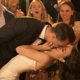 Adrian Drgas ADHD produkcja wideo - Pocałunek pary młodej podczas wesela, w tle goście z zimnymi ogniami. Pan młody w czarnym smokingu, panna młoda w białej sukni. Uśmiechnięci goście weselni.