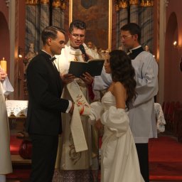 Adrian Drgas ADHD produkcja wideo - Uroczystość ślubna w kościele: para młoda w eleganckich strojach, ksiądz z mikrofonem i książką, asystenci w tle. Dekoracje kwiatowe i czerwony dywan dodają splendoru.