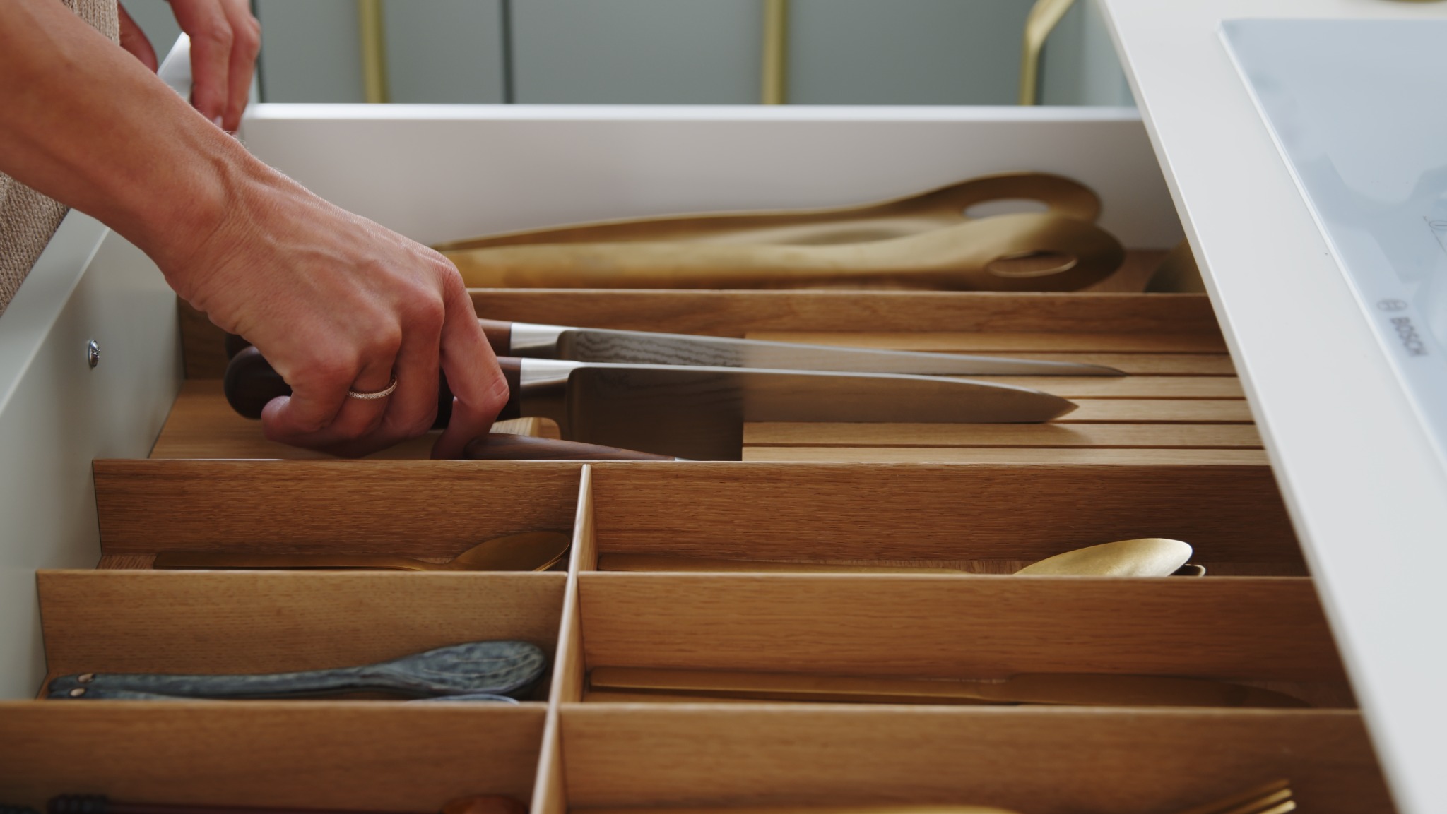 Szczegółowe ujęcie otwartej szuflady z drewnianym organizerem na sztućce i noże; widoczne noże z ciemnymi rękojeściami, złote łyżki i szpatułki, dłoń z pierścionkiem sięgająca do szuflady.