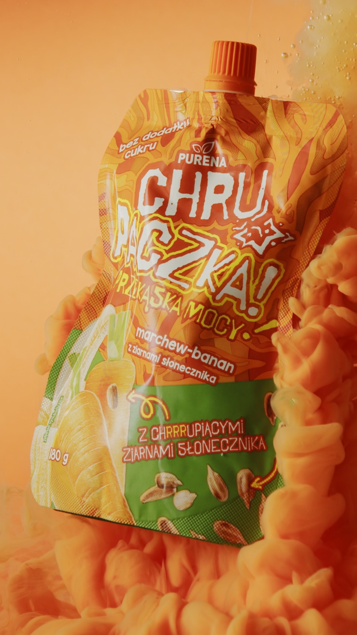 Pionowe ujęcie pomarańczowej saszetki z puree owocowo-warzywnego Purena, zanurzonej w pomarańczowej cieczy przypominającej dym. Saszetka ma nadrukowane grafiki marchewki, banana i ziaren słonecznika.