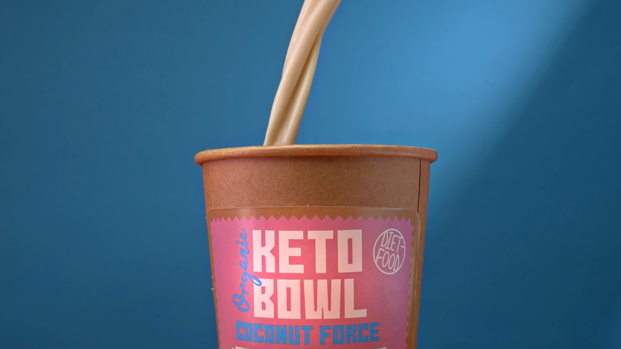 Strumień beżowego płynu wlewa się do kartonowego kubka z etykietą 'Keto Bowl Coconut Force' na tle jednolitego, niebieskiego tła.
