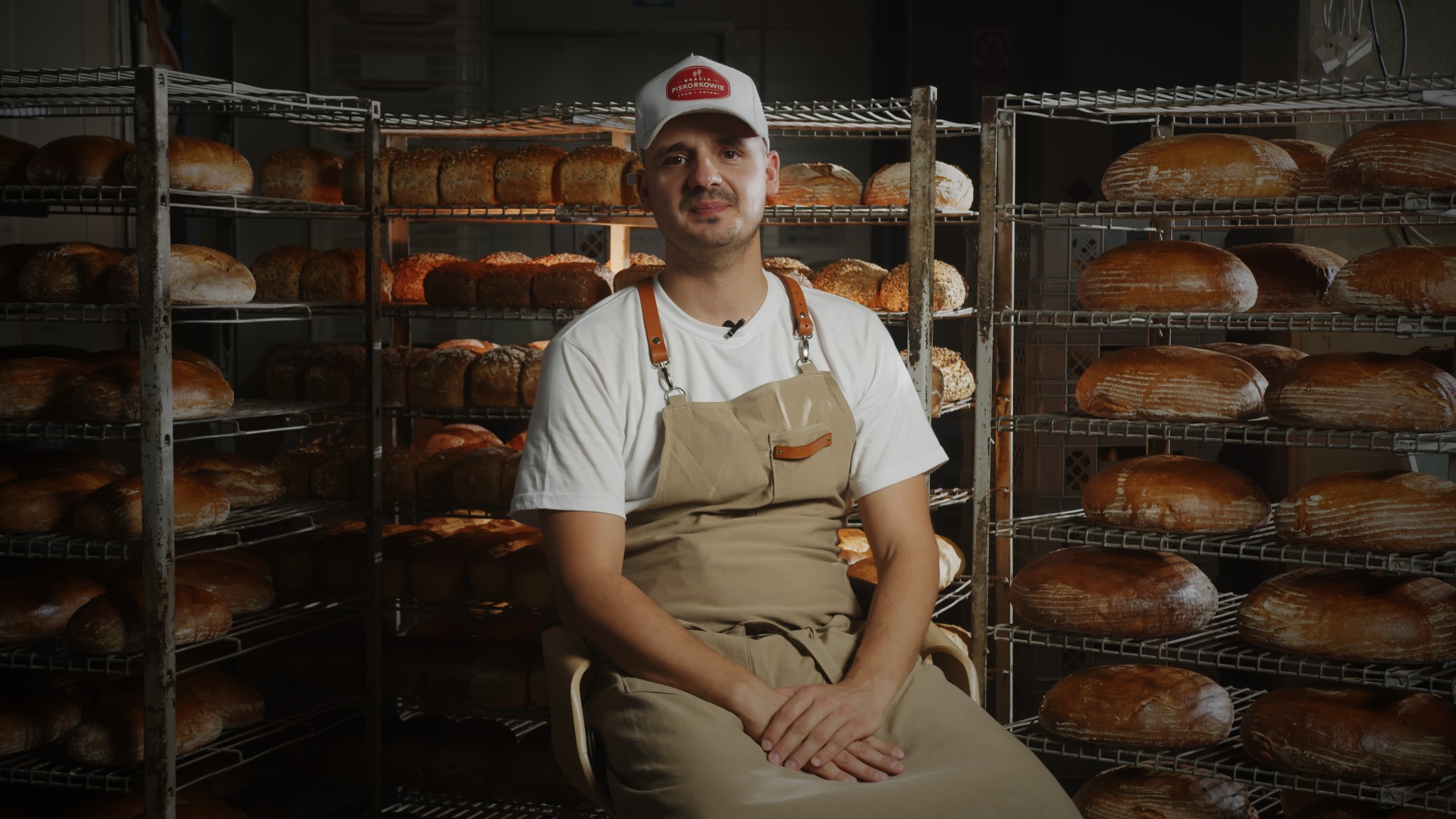 Piekarz siedzi na krześle w fartuchu, na tle regałów z różnym pieczywem, w czapce z logo piekarni.