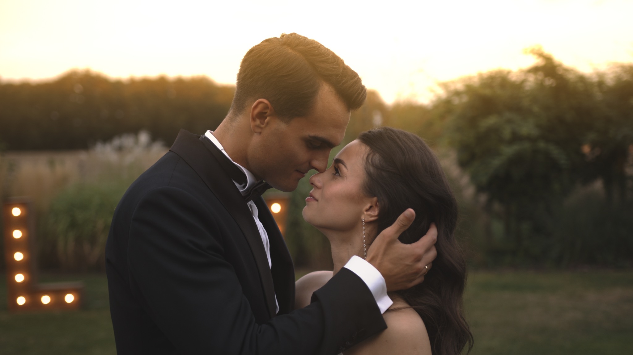 Elegancka para w zbliżeniu, on w smokingu, ona z delikatną biżuterią, w tle dekoracja świetlna i zieleń. Romantyczna atmosfera, ciepłe światło zachodzącego słońca.