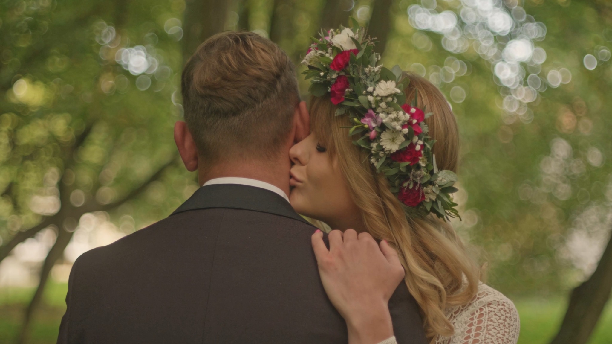 Panna młoda w wianku z czerwonych i białych kwiatów całuje pana młodego w szyję na tle zieleni. Ujęcie z bliska, skupione na parze.