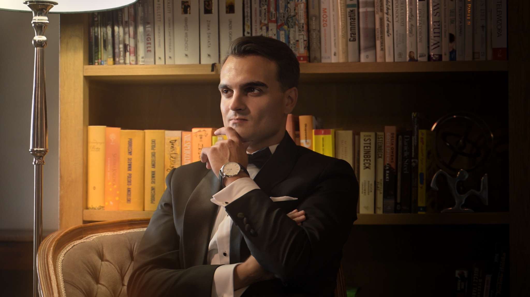 Elegancki mężczyzna w smokingu, z zegarkiem na ręku, siedzi na fotelu na tle regału z książkami, w zamyślonej pozie. Stonowana kolorystyka, miękkie światło.