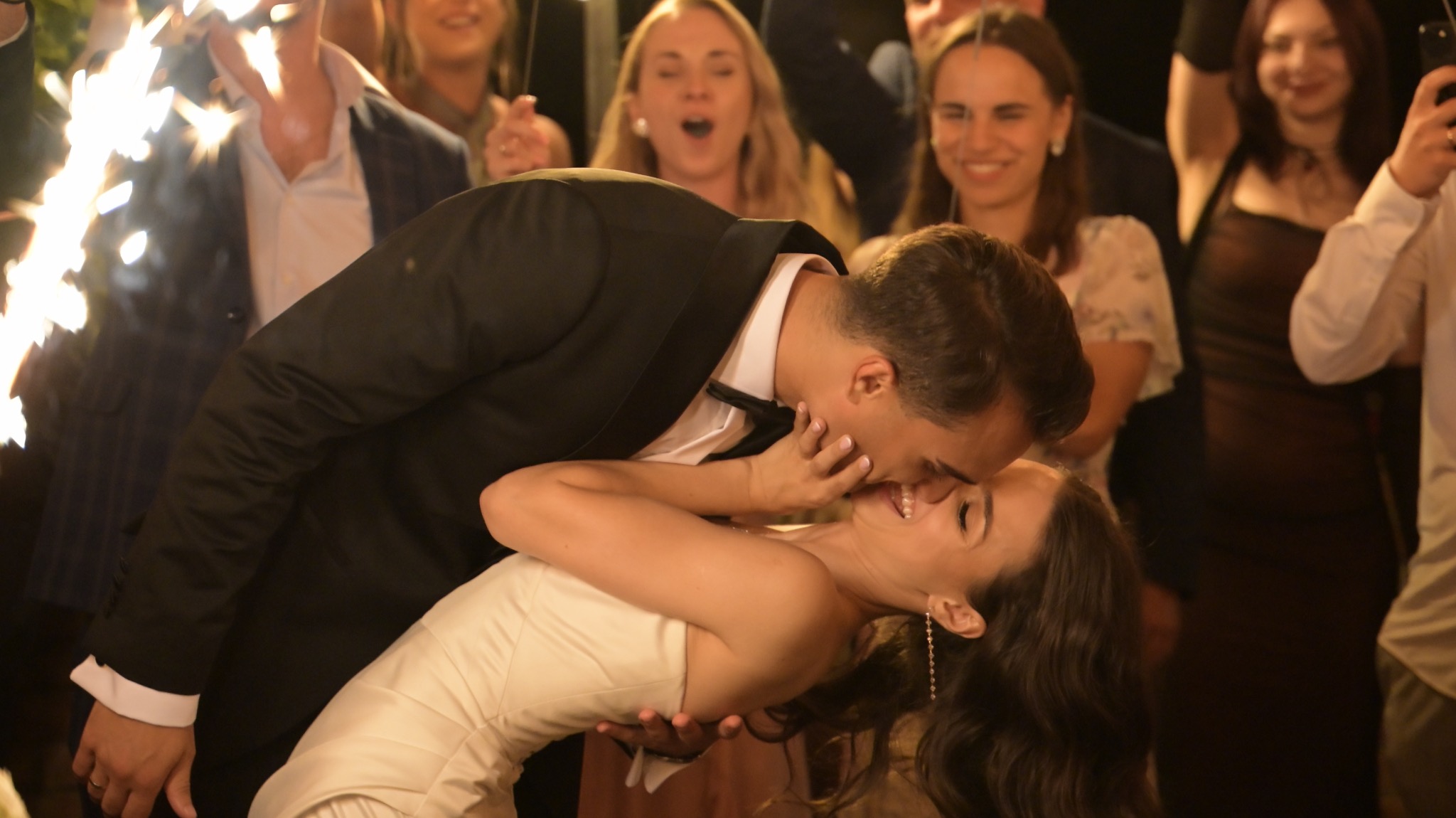 Pocałunek pary młodej podczas wesela, w tle goście z zimnymi ogniami. Pan młody w czarnym smokingu, panna młoda w białej sukni. Uśmiechnięci goście weselni.