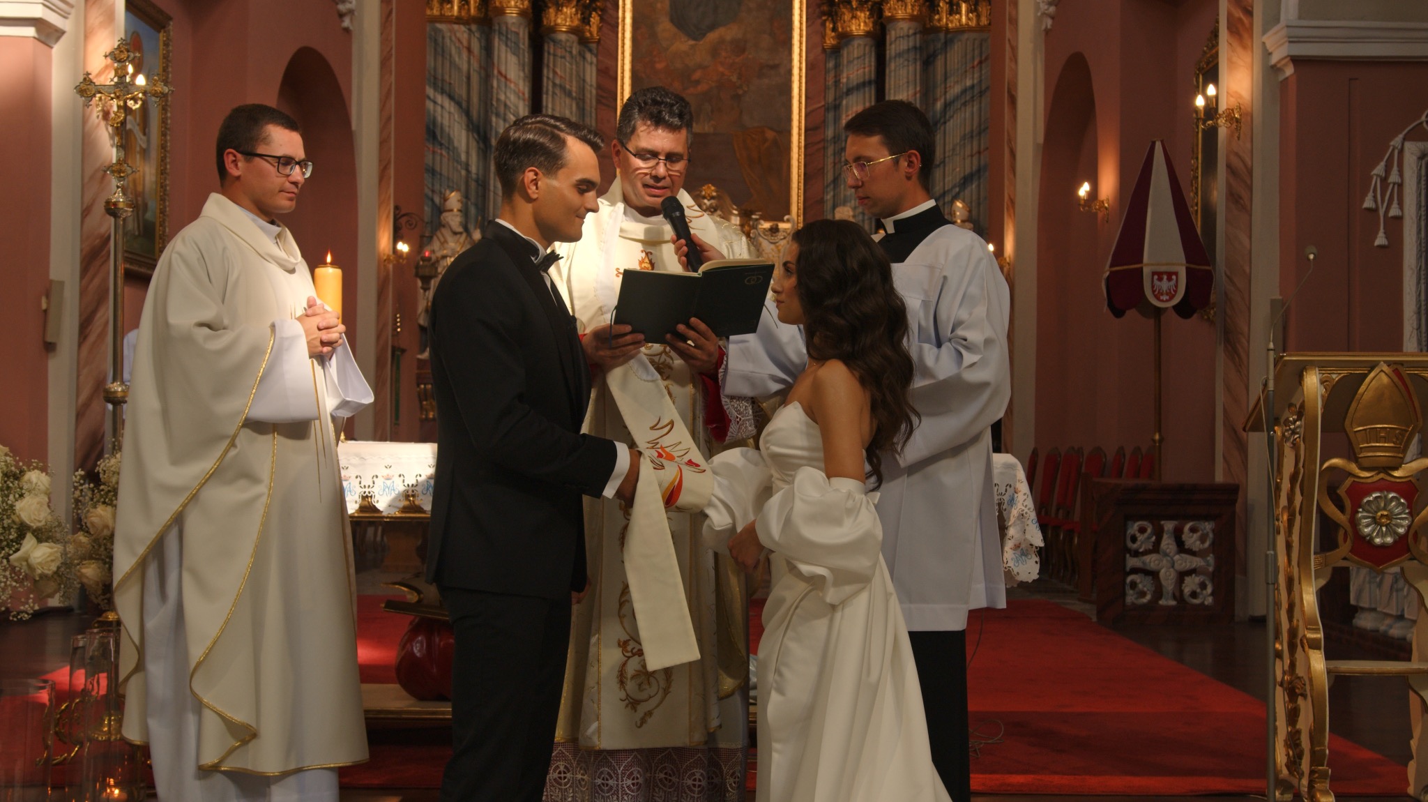 Uroczystość ślubna w kościele: para młoda w eleganckich strojach, ksiądz z mikrofonem i książką, asystenci w tle. Dekoracje kwiatowe i czerwony dywan dodają splendoru.
