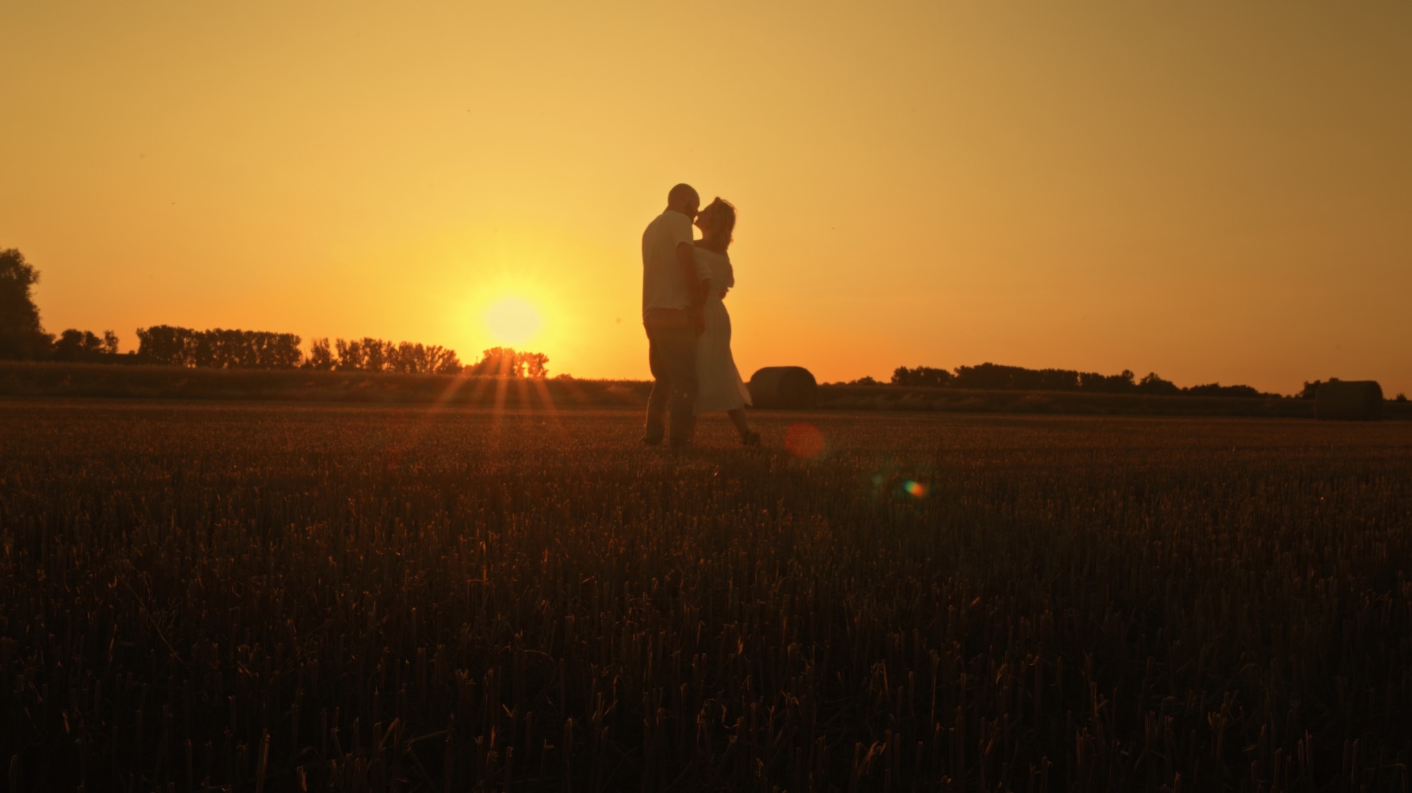 Zakochana para na tle zachodzącego słońca na polu. Silne, ciepłe światło podkreśla romantyczny nastrój. W tle widoczne drzewa i stóg siana.