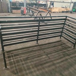 Konstrukcje STAL-HALSBUD - Surowa, metalowa konstrukcja ogrodzenia panelowego z poziomych profili, spawana w warsztacie, widoczna spawarka w tle.