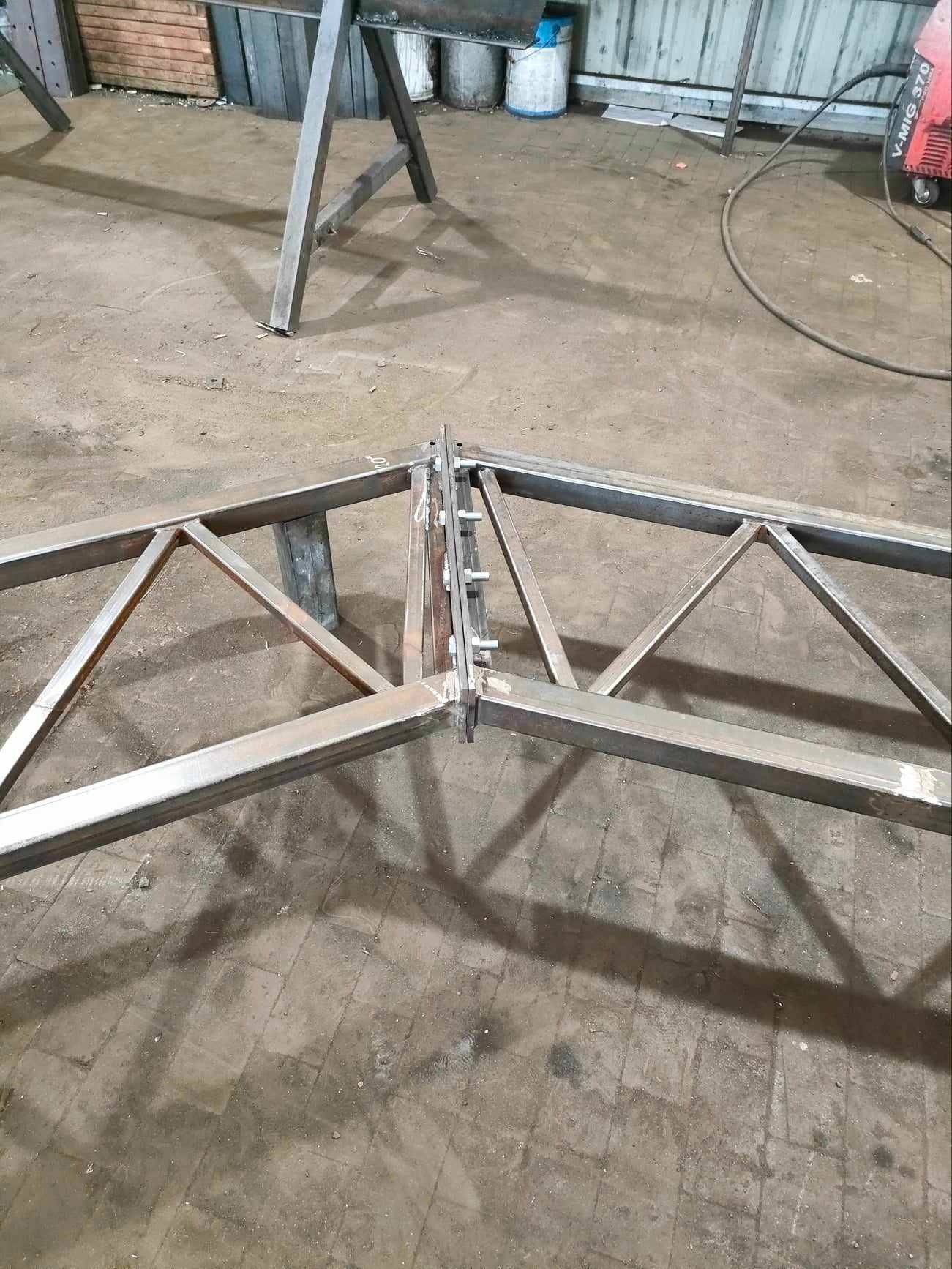 Metalowa konstrukcja kratowa, łączona na śruby, leżąca na podłodze hali warsztatowej, w tle spawarka V-MIG 370