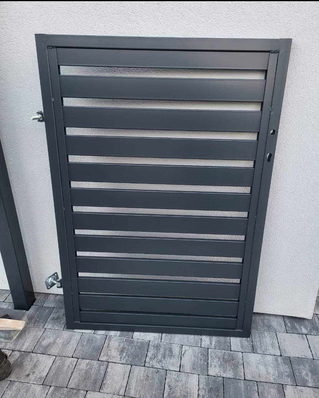 Nowoczesna, szara furtka z poziomymi lamelami, zamontowana na tle jasnoszarej ściany, widoczny fragment kostki brukowej.